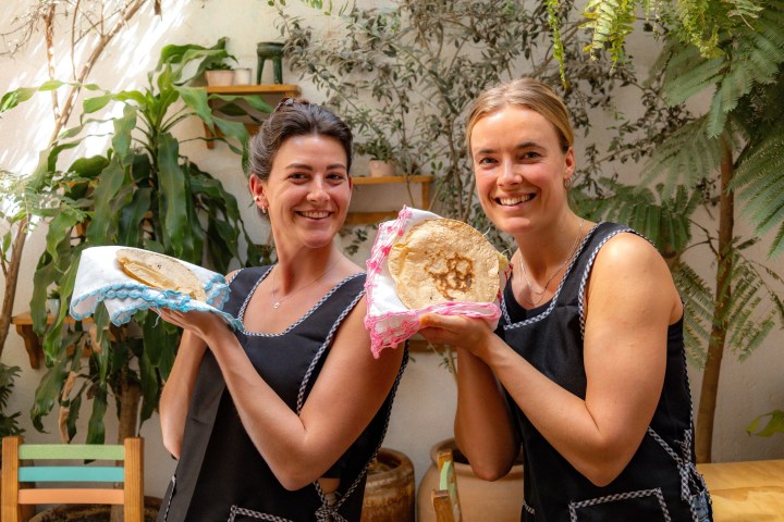 Two smiling women holding flatbreads in a leafy outdoor setting.