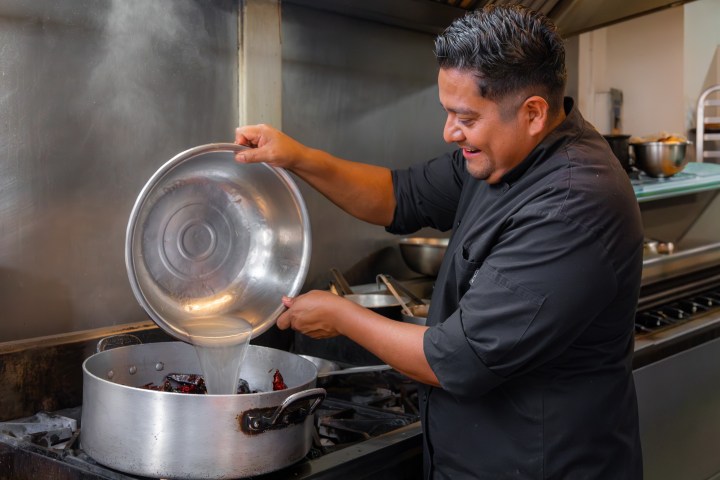 Chef pouring liquid into a pot on a stove in a professional kitchen.