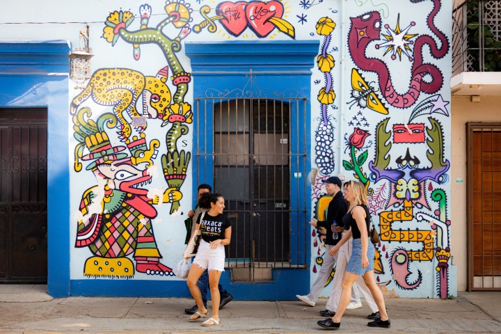 People walk past colorful street art with whimsical characters and patterns on a building wall.