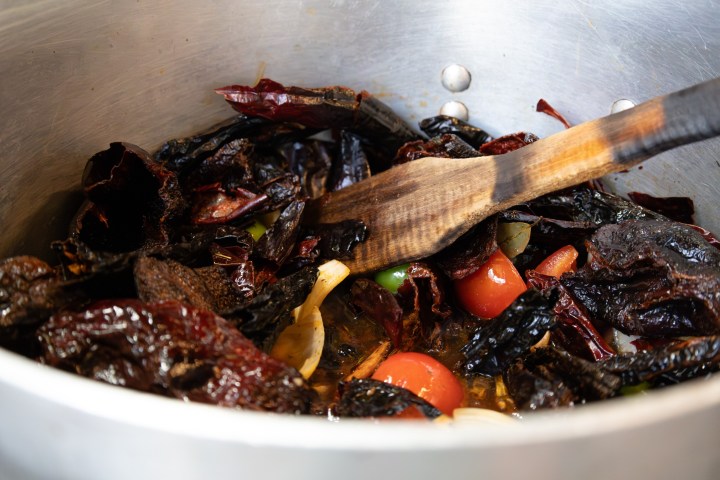 Dried chiles, tomatoes, and onions cooking in a pot with a wooden spatula.