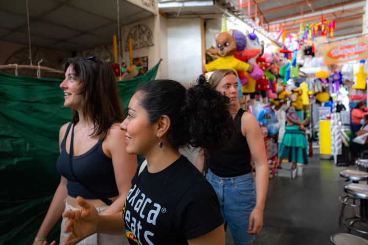 Three women in a market with colorful toys and clothes around.